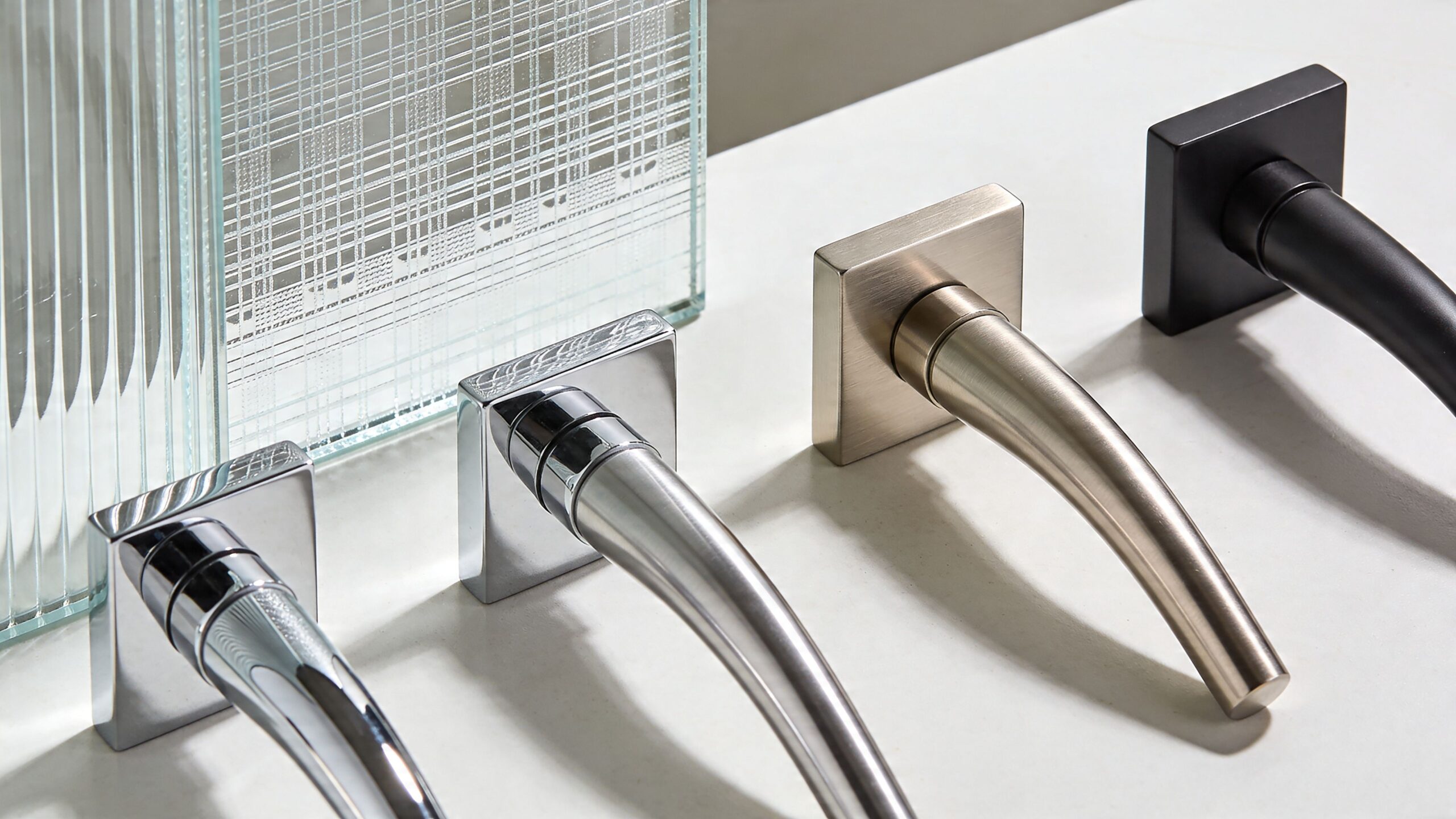 Three different metal door handle designs displayed on a light surface beside a textured glass panel.