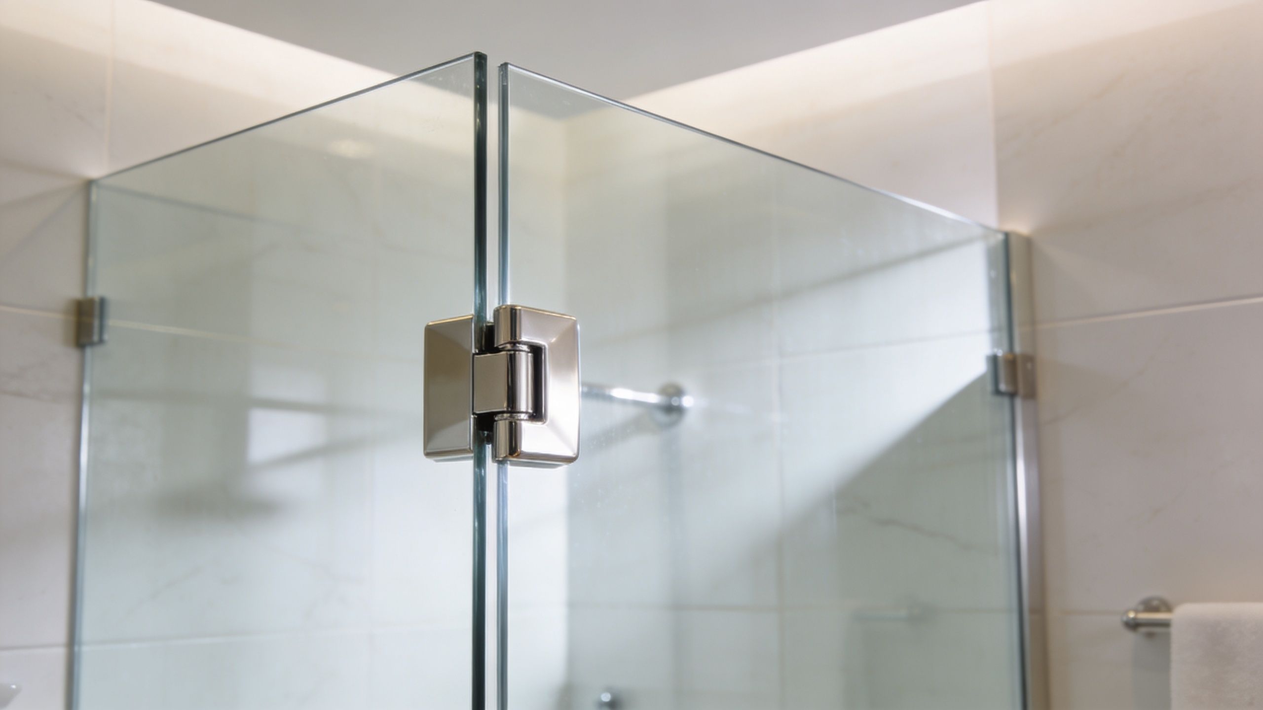 A close-up of chrome glass shower door hinges connecting two glass panels in a modern tiled bathroom.
