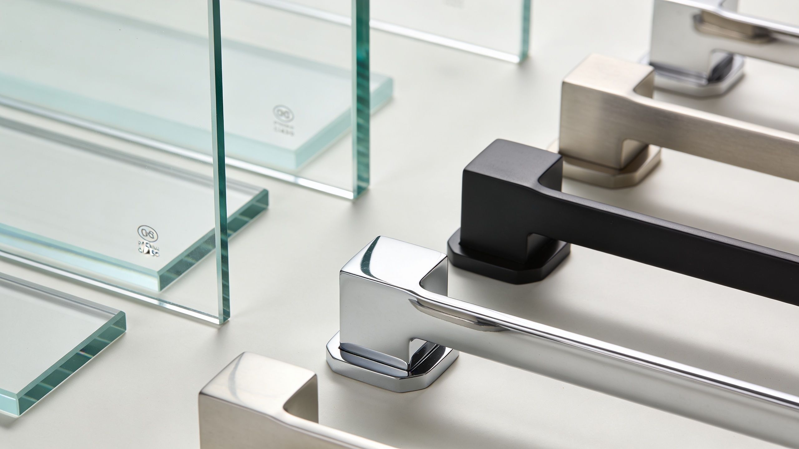 A display of square-based metal shower door handles in various finishes placed next to thick glass panels.